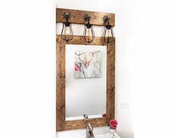 May include: A rectangular wall mirror with a dark brown stained wood frame, featuring an attached top wooden panel holding three black metal cage light fixtures with exposed Edison-style bulbs, reflecting a deer painting.