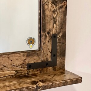 May include: A rustic wooden mirror with a shelf, featuring a dark brown stain and black metal brackets. The mirror has a beveled edge and is framed with reclaimed wood.