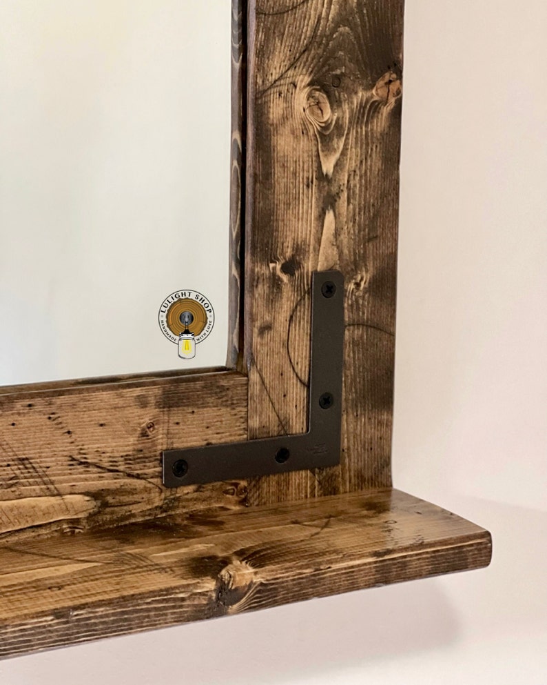May include: A rustic wooden mirror with a shelf, featuring a dark brown stain and black metal brackets. The mirror has a beveled edge and is framed with reclaimed wood.