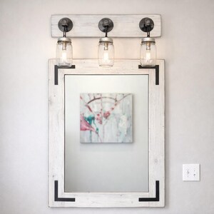 Handcrafted wood framed mirror mounted on a bathroom vanity wall with a three mason jar vanity light fixture on top, featuring a whitewash distressed finish and natural wood grain for a farmhouse-style charm
