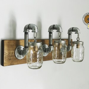 May include: Three glass mason jars with metal lids are suspended from a rustic wooden shelf by industrial pipe fittings. The jars are wired for light fixtures. The shelf is mounted to a white wall. The logo "LULIGHT SHOP" is visible in the upper right corner of the image.