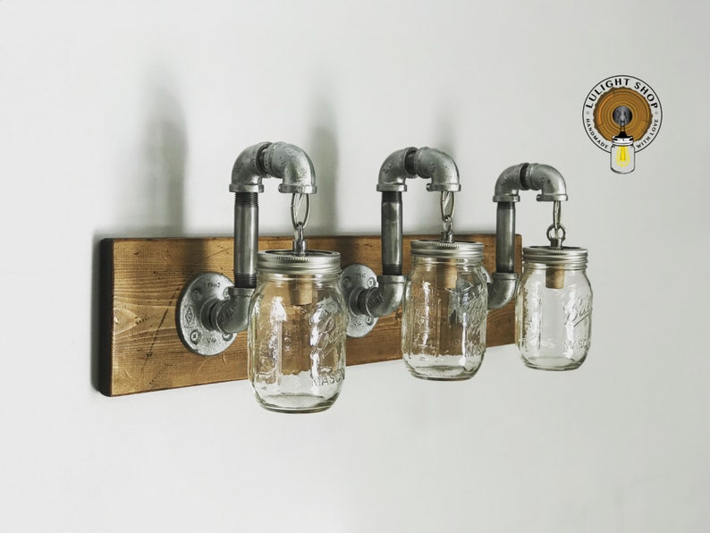 May include: Three glass mason jars with metal lids are suspended from a rustic wooden shelf by industrial pipe fittings. The jars are wired for light fixtures. The shelf is mounted to a white wall. The logo "LULIGHT SHOP" is visible in the upper right corner of the image.