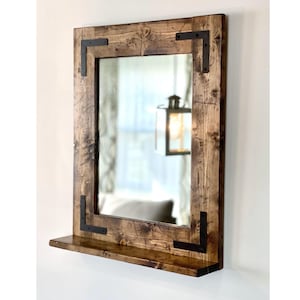 May include: A rectangular wooden mirror with a shelf. The frame is made of stained wood with visible grain and dark metal corner brackets. The mirror reflects a room with a light fixture.