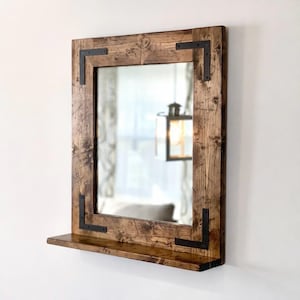 May include: A rustic wooden mirror with a rectangular frame and with a bottom shelf. The frame is made of reclaimed wood and has a dark brown stain. The mirror has a simple, modern design and would be a great addition to any home.