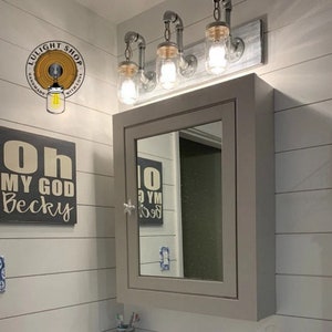 May include: A bathroom vanity with a gray cabinet and a mirror. The cabinet has a star-shaped knob. Above the vanity is a light fixture with three mason jar lights. The light fixture is made of industrial pipes and has a rustic wood base. The wall behind the vanity is white with horizontal wood planks.