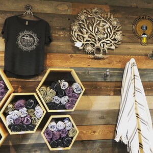 May include: A wooden wall with a black t-shirt with a white graphic design that says "Go Far With My Santa Ynez CA". There are six hexagonal shelves with rolled up fabric in various colors. A white and brown striped towel hangs on a hook. A metal tree-shaped wall decor with flowers and birds is on the wall. A sign that says "LULIGHT SHOP" is on the wall.