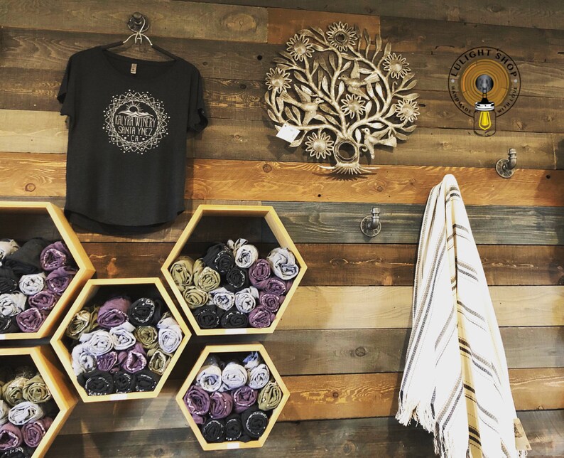 May include: A wooden wall with a black t-shirt with a white graphic design that says "Go Far With My Santa Ynez CA". There are six hexagonal shelves with rolled up fabric in various colors. A white and brown striped towel hangs on a hook. A metal tree-shaped wall decor with flowers and birds is on the wall. A sign that says "LULIGHT SHOP" is on the wall.