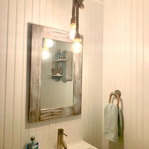 Pu&ograve; includere: Un bagno a tema nautico. Un grande specchio con cornice in legno invecchiato &egrave; appeso sopra un lavandino bianco. Un lampadario a corda con lampadine Edison &egrave; appeso al soffitto. Un portasciugamani con manico in corda contiene un asciugamano azzurro.