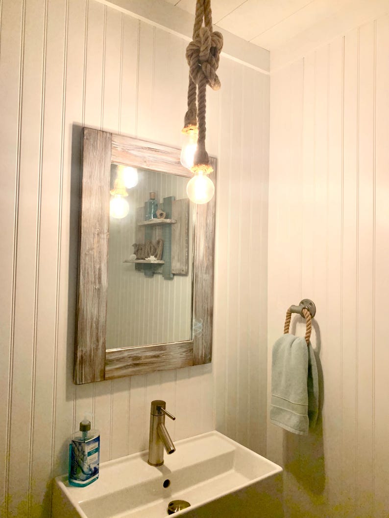 Pu&ograve; includere: Un bagno a tema nautico. Un grande specchio con cornice in legno invecchiato &egrave; appeso sopra un lavandino bianco. Un lampadario a corda con lampadine Edison &egrave; appeso al soffitto. Un portasciugamani con manico in corda contiene un asciugamano azzurro.