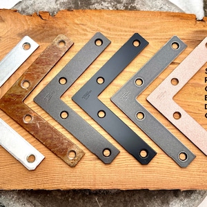 Puede incluir: Seis soportes de esquina de metal en diferentes acabados: plateado, oxidado, bronce frotado con aceite, negro, n&iacute;quel oscuro y cobre. Los soportes est&aacute;n dispuestos en una fila sobre una superficie de madera.