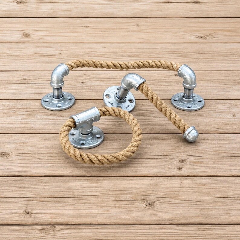 May include: A set of industrial-style bathroom fixtures. The set includes a towel bar, a towel ring, and a toilet paper holder. Each piece features galvanized metal pipe fittings and natural rope. The fixtures are mounted on round metal plates.
