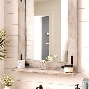 May include: A rectangular, rustic wooden mirror with a shelf, featuring a light gray finish and black metal corner accents. The shelf holds a small candle, a vase with flowers, and a mug. The mirror is mounted on a white, horizontal plank wall.