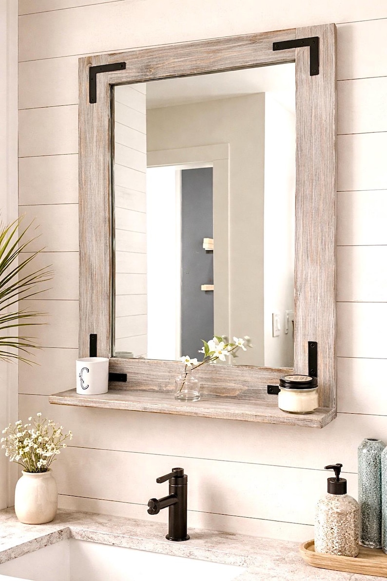 May include: A rectangular, rustic wooden mirror with a shelf, featuring a light gray finish and black metal corner accents. The shelf holds a small candle, a vase with flowers, and a mug. The mirror is mounted on a white, horizontal plank wall.