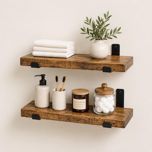May include: Two rustic wooden shelves with black metal brackets are mounted on a white wall. The top shelf holds white towels, a small white vase with greenery, and a black box. The bottom shelf displays a soap dispenser, toothbrushes, a candle, and a jar of cotton balls.