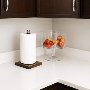 A silver metal pipe paper towel holder with a wooden base sits on a white countertop next to a clear glass bowl filled with red tomatoes and yellow apples.