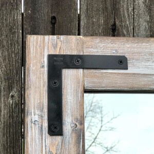 Pu&ograve; includere: Primo piano di una cornice di specchio in legno rustico con finitura bianca anticata. La cornice &egrave; unita agli angoli con staffe a L in metallo nero fissate con viti nere. Lo sfondo &egrave; una staccionata in legno usurato.