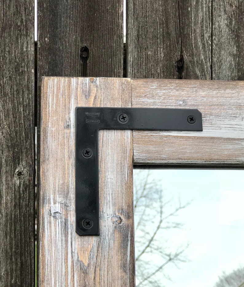 Pu&ograve; includere: Primo piano di una cornice di specchio in legno rustico con finitura bianca anticata. La cornice &egrave; unita agli angoli con staffe a L in metallo nero fissate con viti nere. Lo sfondo &egrave; una staccionata in legno usurato.