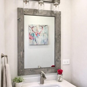 Handcrafted wood framed mirror mounted on a bathroom vanity wall with a three mason jar vanity light fixture on top, featuring a gray finish and natural wood grain for a farmhouse-style charm