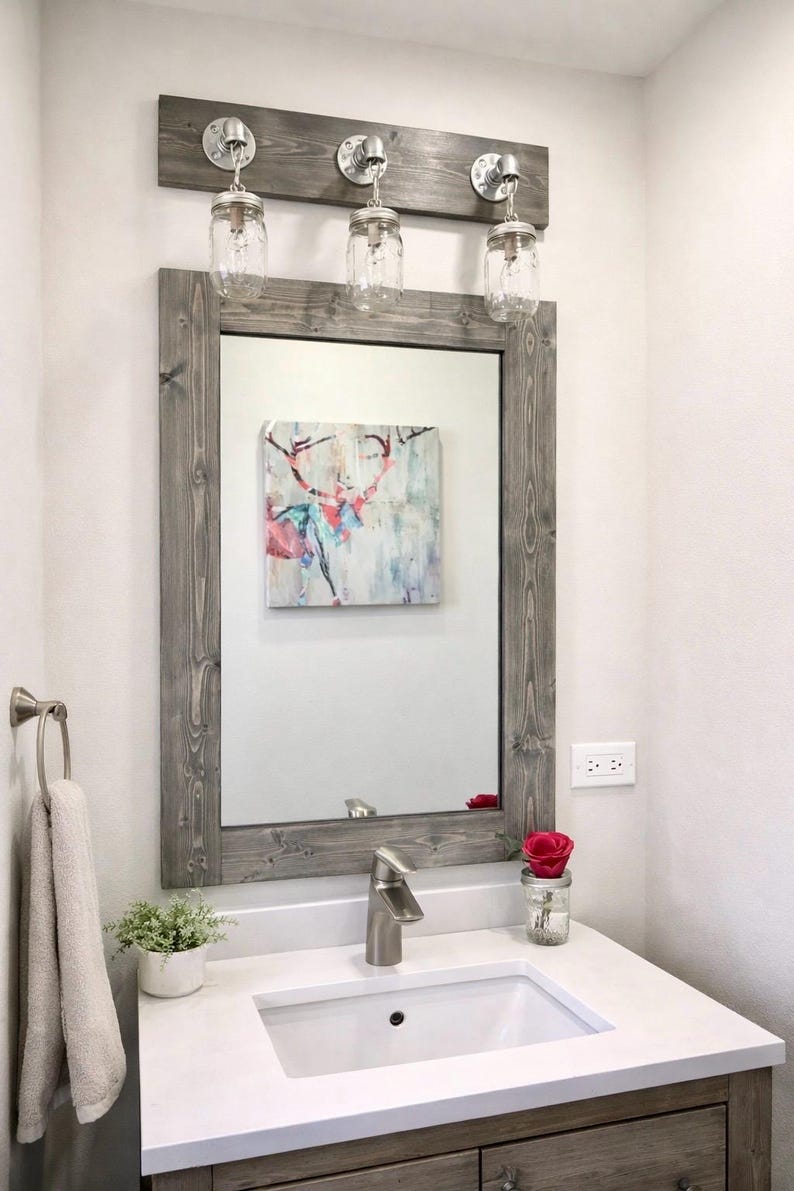Handcrafted wood framed mirror mounted on a bathroom vanity wall with a three mason jar vanity light fixture on top, featuring a gray finish and natural wood grain for a farmhouse-style charm