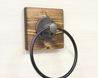 May include: A rustic towel ring made of a dark stained wooden square base with a black metal ring and mounting hardware. The ring is attached to a metal fitting, creating an industrial look. The wood grain is visible.