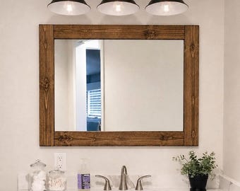 May include: A rectangular wall mirror with a dark brown stained wooden frame, positioned above a bathroom vanity. Above the mirror is a three-light fixture with black metal hardware and white dome-shaped shades, providing downward illumination.