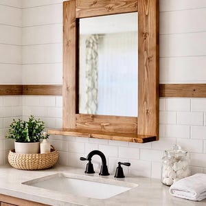 May include: A rectangular wooden framed mirror with a shelf, mounted on a white tiled wall. The mirror is above a white sink with black fixtures. A woven basket with plants and a jar of cotton balls are on the counter.