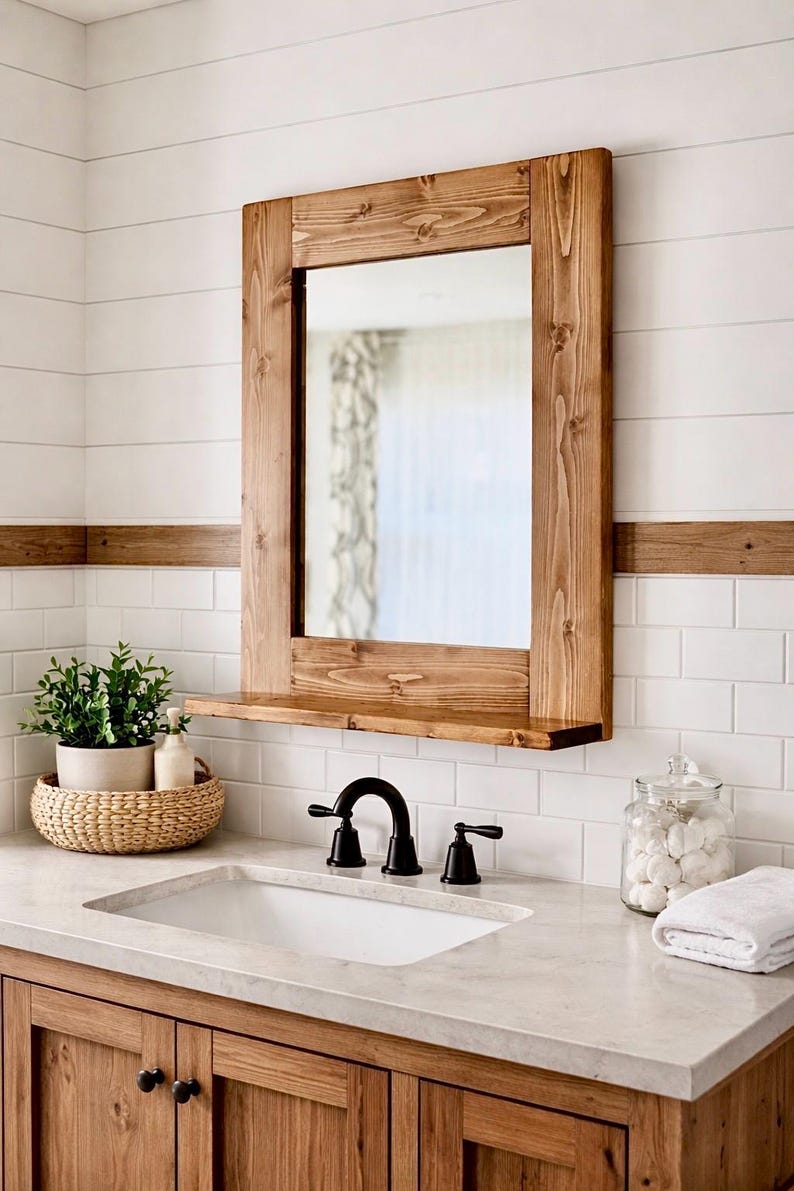 May include: A rectangular wooden framed mirror with a shelf, mounted on a white tiled wall. The mirror is above a white sink with black fixtures. A woven basket with plants and a jar of cotton balls are on the counter.
