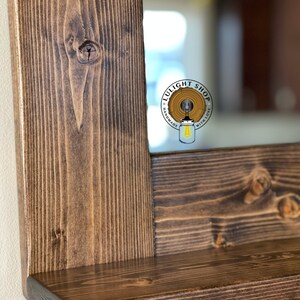 Puede incluir: Primer plano de un marco de espejo rectangular de madera con una peque&ntilde;a repisa. El marco es de madera te&ntilde;ida de color marr&oacute;n oscuro, mostrando la veta natural y los nudos de la madera. El espejo refleja un interior borroso. El logotipo "LULIGHT SHOP" es visible.