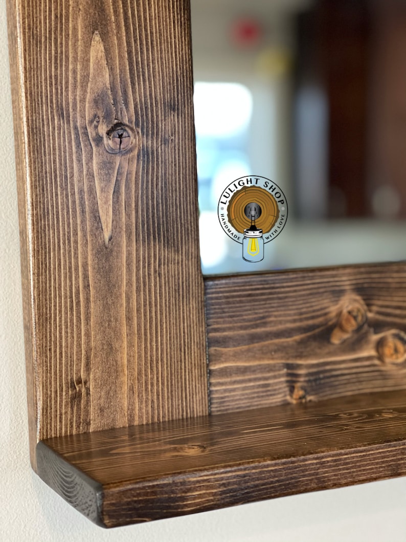 Puede incluir: Primer plano de un marco de espejo rectangular de madera con una peque&ntilde;a repisa. El marco es de madera te&ntilde;ida de color marr&oacute;n oscuro, mostrando la veta natural y los nudos de la madera. El espejo refleja un interior borroso. El logotipo "LULIGHT SHOP" es visible.