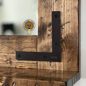 May include: A rustic wooden framed mirror with a shelf. The frame is made of dark stained wood with visible grain and knots. Dark metal L-brackets secure the shelf to the frame. The mirror reflects a light-colored background.