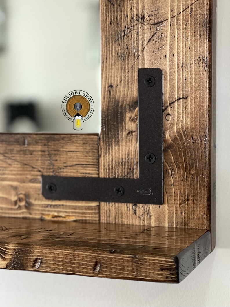 May include: A rustic wooden framed mirror with a shelf. The frame is made of dark stained wood with visible grain and knots. Dark metal L-brackets secure the shelf to the frame. The mirror reflects a light-colored background.