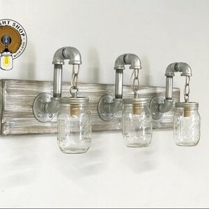 May include: Three mason jar light fixtures with metal pipes and a wooden base. The jars are clear glass and have a rustic look. The base is a light wood with a whitewashed finish.