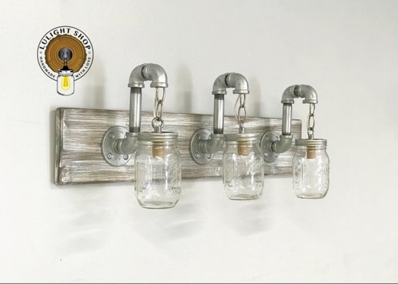 May include: Three mason jar light fixtures with metal pipes and a wooden base. The jars are clear glass and have a rustic look. The base is a light wood with a whitewashed finish.