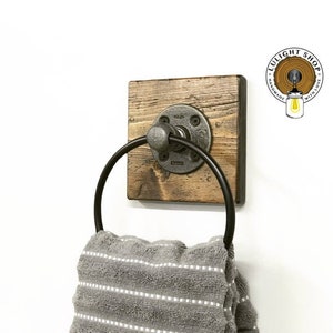 May include: A rustic wooden towel ring with a black metal ring and a pipe fitting. The ring is mounted on a square piece of wood with a distressed finish. The towel ring is hanging on a white wall.