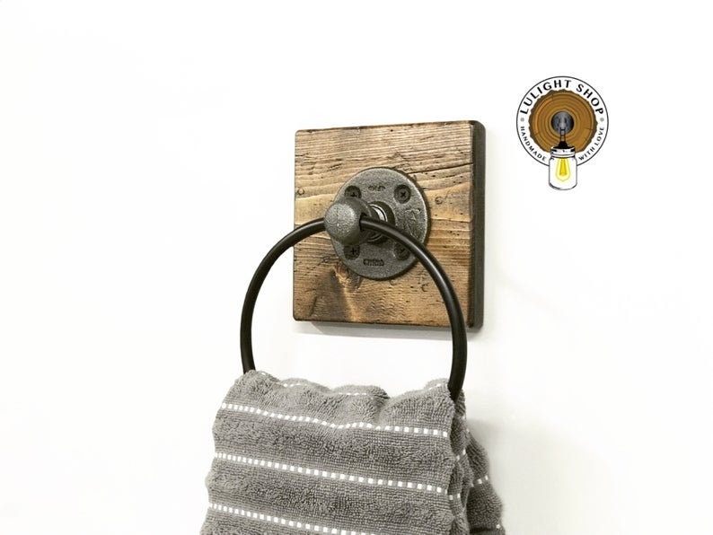 May include: A rustic wooden towel ring with a black metal ring and a pipe fitting. The ring is mounted on a square piece of wood with a distressed finish. The towel ring is hanging on a white wall.