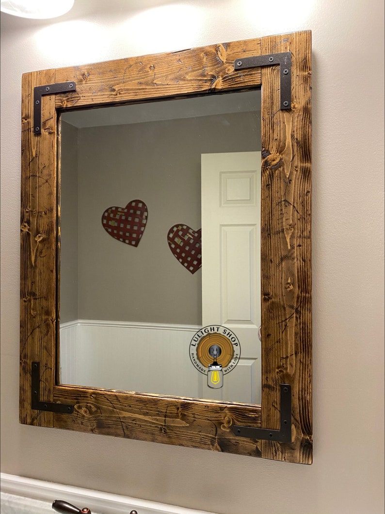 Puede incluir: Un espejo de madera r&uacute;stico con una mancha marr&oacute;n oscura y soportes de esquina de metal negro. El espejo es rectangular y tiene un dise&ntilde;o simple. El espejo refleja un ba&ntilde;o con paredes blancas y una puerta blanca.