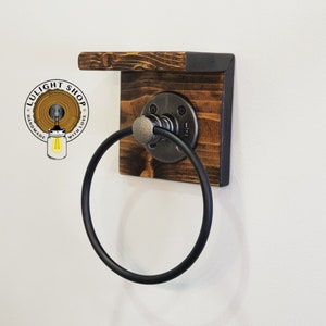 May include: A black metal towel ring attached to a wooden shelf with a black metal frame. The shelf is made of reclaimed wood and has a rustic look. The towel ring is mounted to the shelf with a metal bracket.