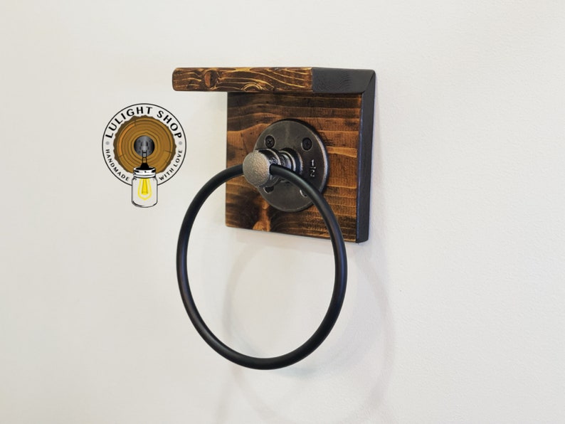 May include: A black metal towel ring attached to a wooden shelf with a black metal frame. The shelf is made of reclaimed wood and has a rustic look. The towel ring is mounted to the shelf with a metal bracket.