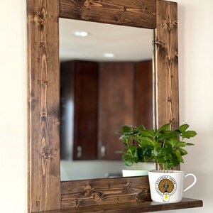 Puede incluir: Espejo rectangular de madera con marco te&ntilde;ido de color marr&oacute;n oscuro y una peque&ntilde;a repisa. Una taza blanca con una planta y el texto "LULIGHT SHOP" se encuentra en la repisa. El espejo refleja una habitaci&oacute;n con armarios marrones.