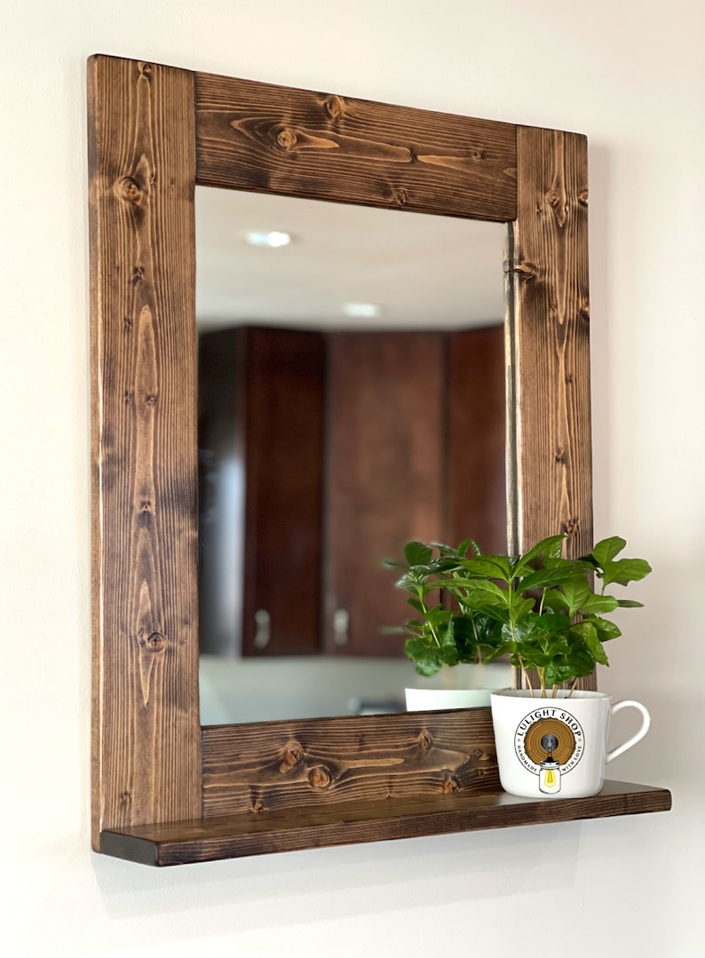 Puede incluir: Espejo rectangular de madera con marco te&ntilde;ido de color marr&oacute;n oscuro y una peque&ntilde;a repisa. Una taza blanca con una planta y el texto "LULIGHT SHOP" se encuentra en la repisa. El espejo refleja una habitaci&oacute;n con armarios marrones.