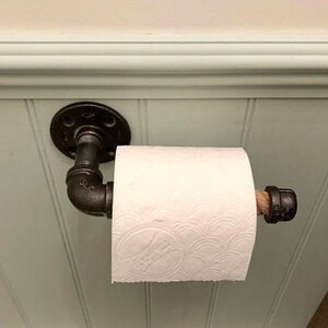 May include: Industrial-style toilet paper holder made from black metal pipes. The holder is mounted on a light green wall and holds a roll of white toilet paper. The design incorporates a rustic aesthetic.