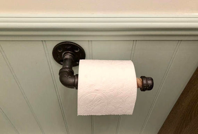 May include: Industrial-style toilet paper holder made from black metal pipes. The holder is mounted on a light green wall and holds a roll of white toilet paper. The design incorporates a rustic aesthetic.
