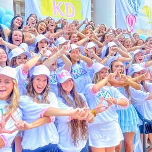 May include: A group of people wearing white t-shirts with pink and blue lettering, white baseball caps, and skirts or shorts. The shirts feature the text KD and KA. Large banners with the text KD and hot air balloon illustrations hang in the background.