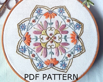 May include: A white embroidery hoop with a floral mandala design. The design features pink, orange, and blue flowers with gold accents. The text "PDF PATTERN" is printed on the hoop.