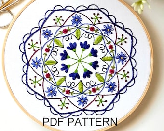 May include: Embroidered hoop art with a floral mandala design. The design features blue, green, red, and white flowers on a white background. The hoop is made of wood and has a diameter of approximately 8 inches. The text "PDF PATTERN" is printed on the hoop.