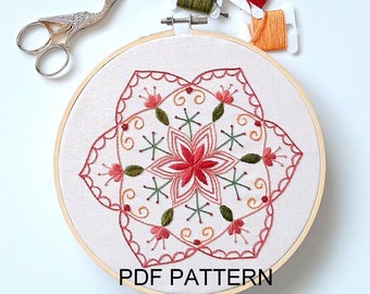 May include: Embroidered floral mandala pattern in a wooden embroidery hoop with red, pink, green, and yellow thread. The hoop is on a white surface with scissors and spools of thread.