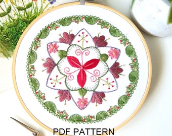 May include: A white embroidery hoop with a floral design in shades of pink, green, and red. The design features a central flower with a starburst pattern and a border of leaves and flowers. The hoop is on a white surface with a pair of scissors and the text "PDF PATTERN".