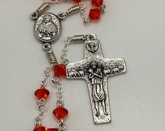 Könnte beinhalten: Ein Rosenkranz mit roten facettierten Perlen und silberfarbenen Metallkomponenten. Das Kreuz zeigt ein detailliertes Relief einer Figur mit Schafen. Der Rosenkranz enthält auch ein Medaillon mit einer religiösen Figur.