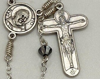 Könnte beinhalten: Ein silberner Rosenkranz mit dunkelgrauen facettierten Perlen. Der Rosenkranz hat ein Kruzifix mit der Inschrift "INRI" und eine runde Medaille mit dem Bild eines Heiligen. Die Kettenglieder sind filigran und das Metall hat eine polierte Oberfläche.