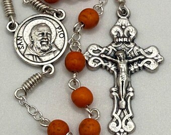 Könnte beinhalten: Ein Rosenkranz mit orangefarbenen Perlen und silberfarbenen Metallkomponenten. Der Rosenkranz hat ein Kruzifix mit einer detaillierten Darstellung Jesu und einen runden Anhänger mit dem Bild eines Heiligen und dem Text "SAN PIO".